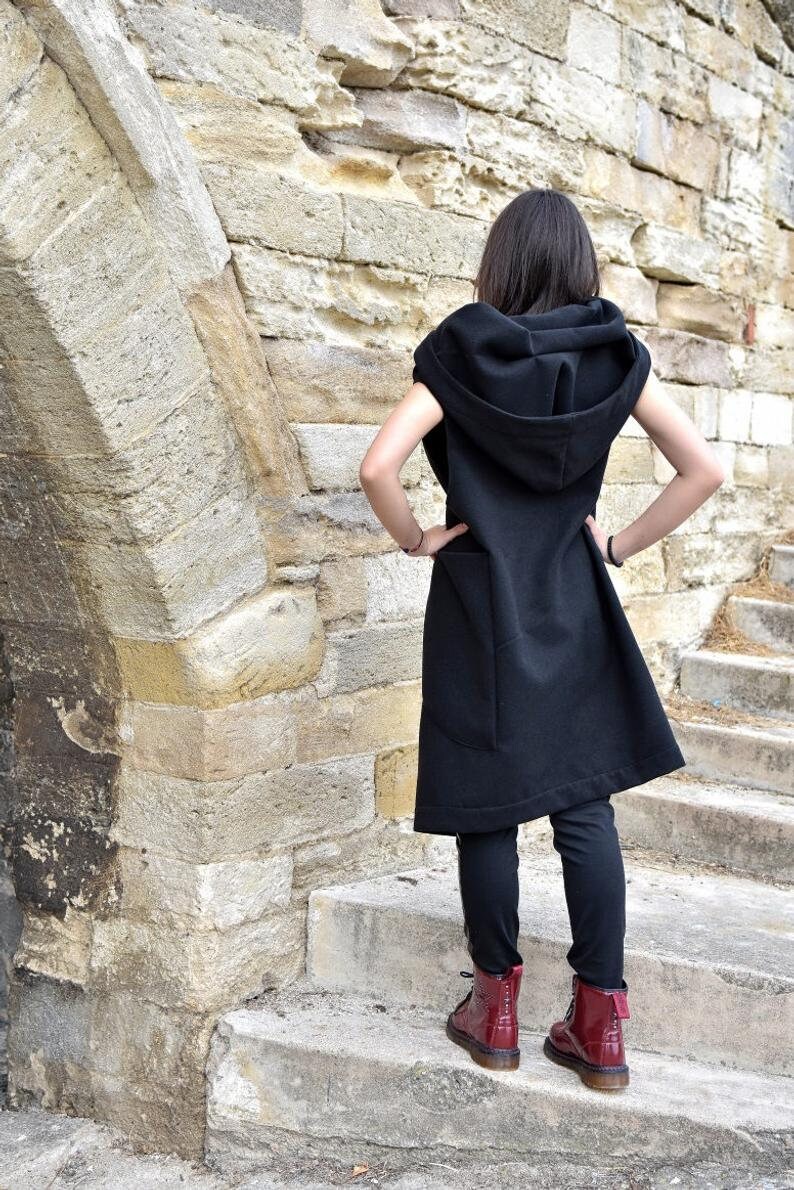 May include: A black sleeveless hooded vest with a long, flowing design. The vest has a large hood and a single pocket on the front. The person wearing the vest is standing on a set of stone steps.