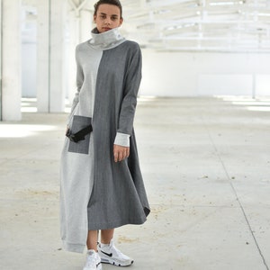 Puede incluir: Un vestido largo de manga larga gris y negro con cuello alto y cintur&oacute;n negro. El vestido tiene un corte holgado y un dobladillo ligeramente asim&eacute;trico. La modelo lleva zapatillas blancas.