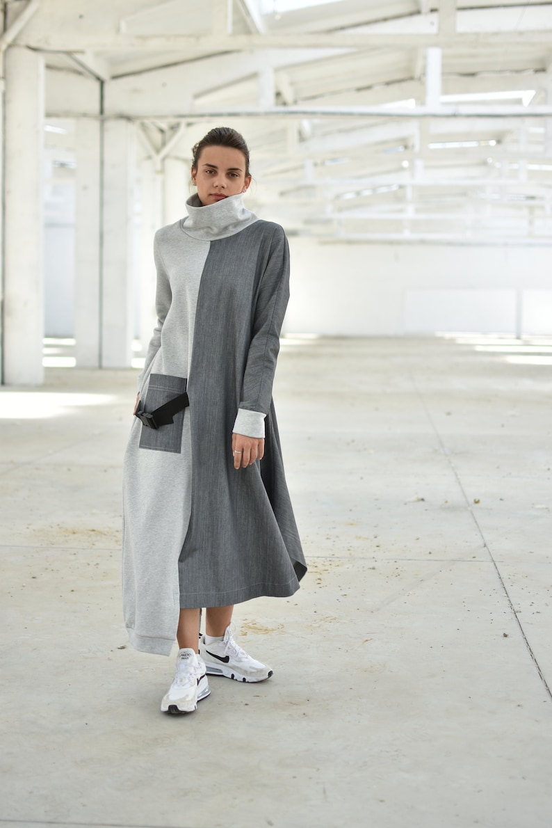 Puede incluir: Un vestido largo de manga larga gris y negro con cuello alto y cintur&oacute;n negro. El vestido tiene un corte holgado y un dobladillo ligeramente asim&eacute;trico. La modelo lleva zapatillas blancas.