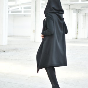 May include: A person wearing a long black hooded coat and black pants. The coat has a flowing design and a large hood. The person is wearing black boots with silver accents. The background is a white industrial space.