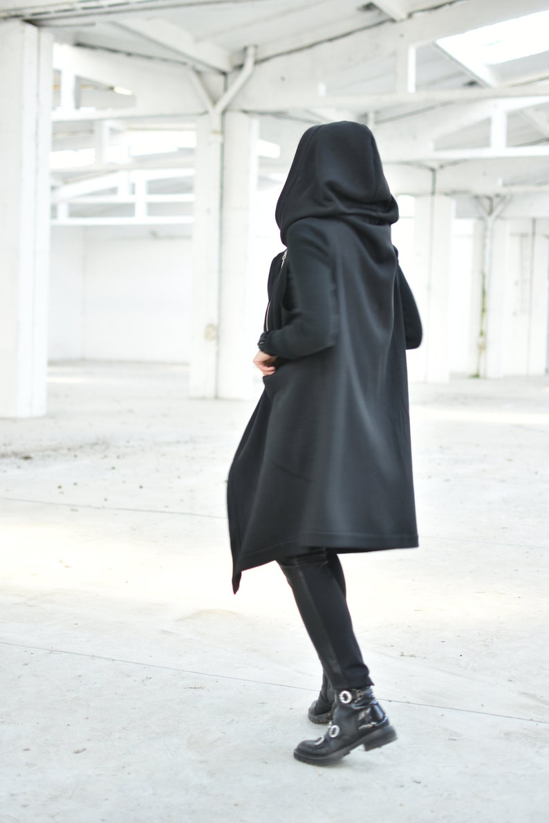 May include: A person wearing a long black hooded coat and black pants. The coat has a flowing design and a large hood. The person is wearing black boots with silver accents. The background is a white industrial space.
