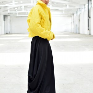 May include: A person wearing a bright yellow, quilted jacket and a long, flowing black skirt. The jacket has a high collar and the skirt drapes to the floor. The outfit is set against a white, industrial background.