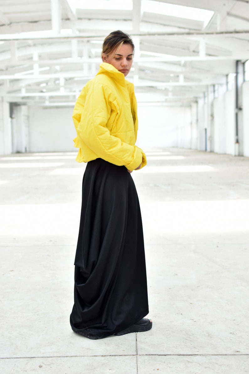 May include: A person wearing a bright yellow, quilted jacket and a long, flowing black skirt. The jacket has a high collar and the skirt drapes to the floor. The outfit is set against a white, industrial background.