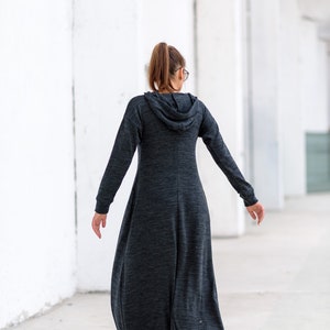 May include: A woman wearing a long, dark gray hooded dress with a relaxed fit. The dress has a long, flowing skirt and a drawstring hood.