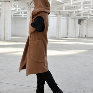 May include: A long brown hooded vest with a large pocket on the front. The vest is worn over a black shirt and black trousers.