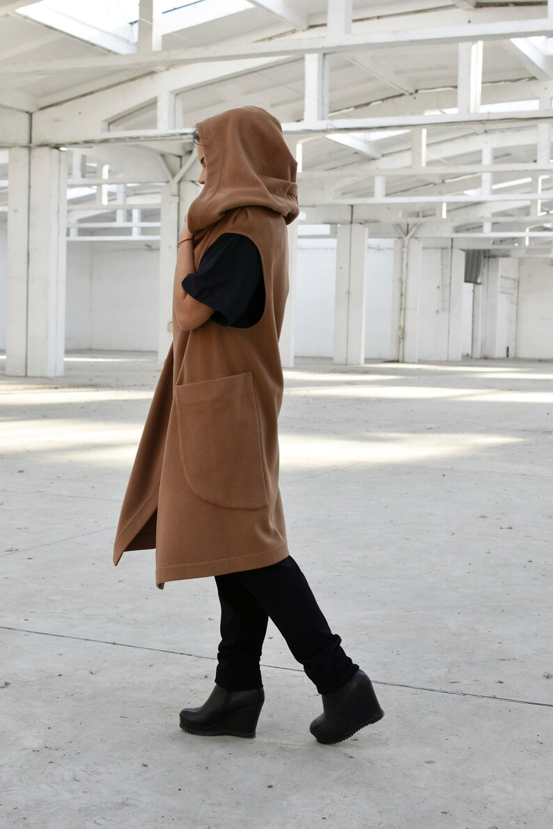 May include: A long brown hooded vest with a large pocket on the front. The vest is worn over a black shirt and black trousers.