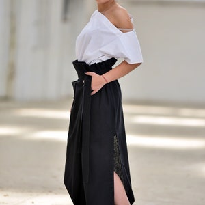 May include: A woman wearing a white off-the-shoulder top and a black maxi skirt with a high waist and a side slit. The skirt has a wide black belt with a buckle. She is wearing white sneakers.