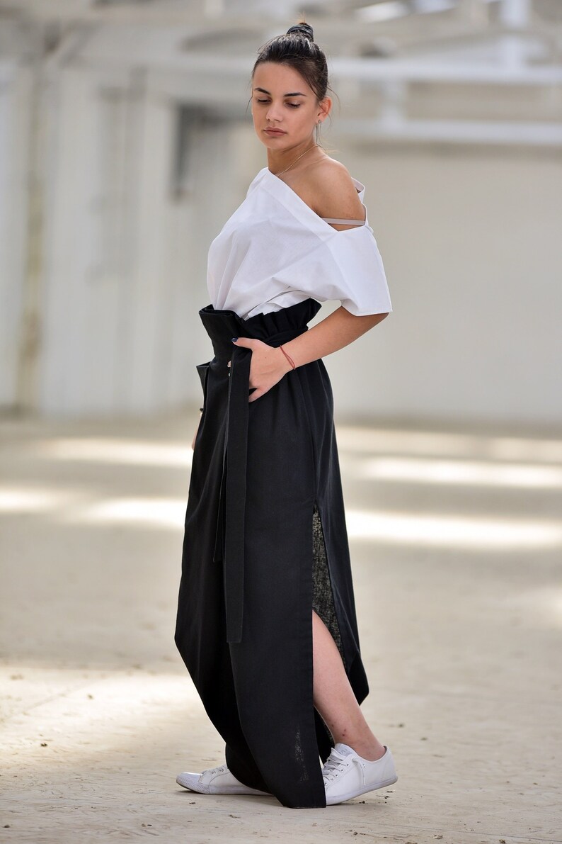 May include: A woman wearing a white off-the-shoulder top and a black maxi skirt with a high waist and a side slit. The skirt has a wide black belt with a buckle. She is wearing white sneakers.