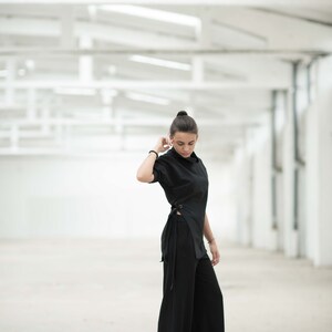 May include: A woman wearing a black long-sleeved top with a side tie and black wide-leg trousers. She is standing in a white room with a white ceiling.