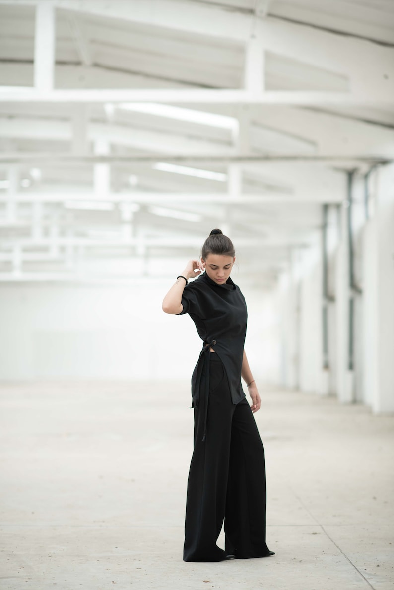 May include: A woman wearing a black long-sleeved top with a side tie and black wide-leg trousers. She is standing in a white room with a white ceiling.