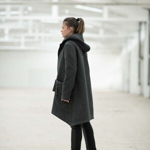May include: A woman wearing a gray hooded coat with a long, asymmetrical hemline. She is wearing black pants and black wedge boots.