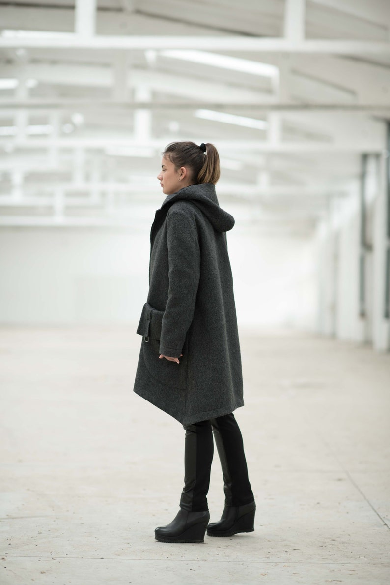 May include: A woman wearing a gray hooded coat with a long, asymmetrical hemline. She is wearing black pants and black wedge boots.
