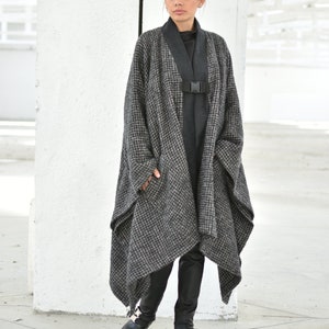 Pu&ograve; includere: Un lungo cappotto a quadri bianco e nero con una cintura e fibbia nere. Il cappotto &egrave; indossato sopra un dolcevita nero e pantaloni neri. La persona indossa scarpe nere con fibbie argentate.
