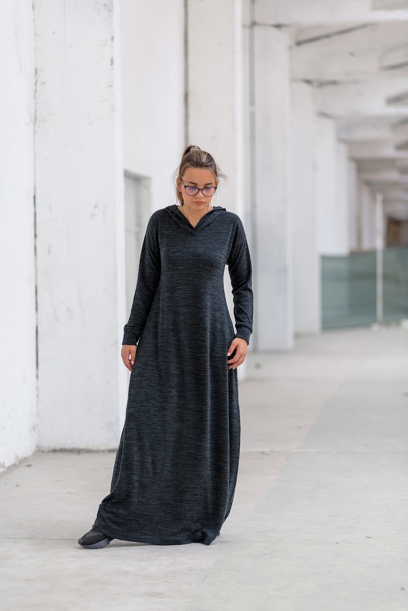 May include: A woman wearing a long, dark gray hooded dress with a relaxed fit. The dress has a slightly textured fabric and long sleeves. She is standing in a white hallway.