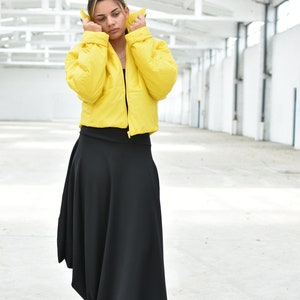 May include: A woman wearing a bright yellow puffer jacket and a long black skirt. The jacket is cropped and the skirt has an asymmetrical hemline. The outfit is set against a white background.