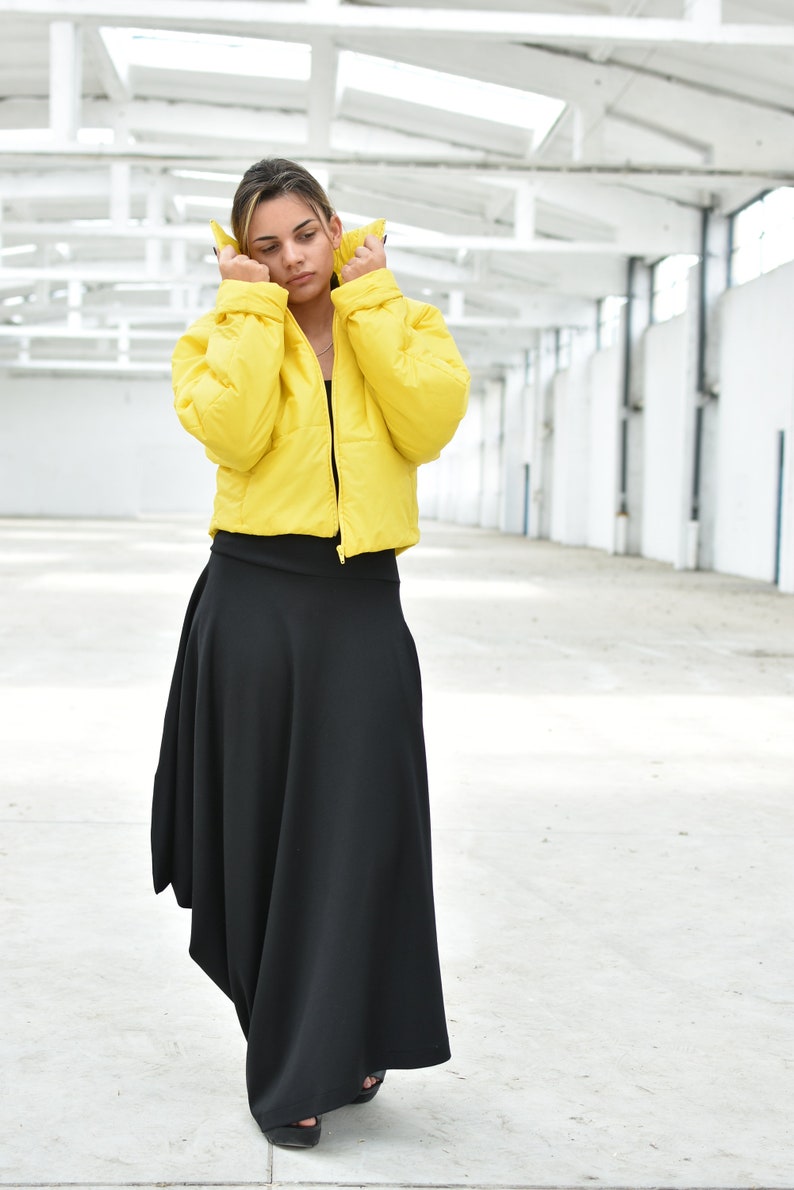 May include: A woman wearing a bright yellow puffer jacket and a long black skirt. The jacket is cropped and the skirt has an asymmetrical hemline. The outfit is set against a white background.
