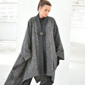 Pu&ograve; includere: Un poncho a motivi grigi e neri con una chiusura a fibbia nera. Il poncho ha un orlo lungo e asimmetrico ed &egrave; indossato sopra pantaloni neri.