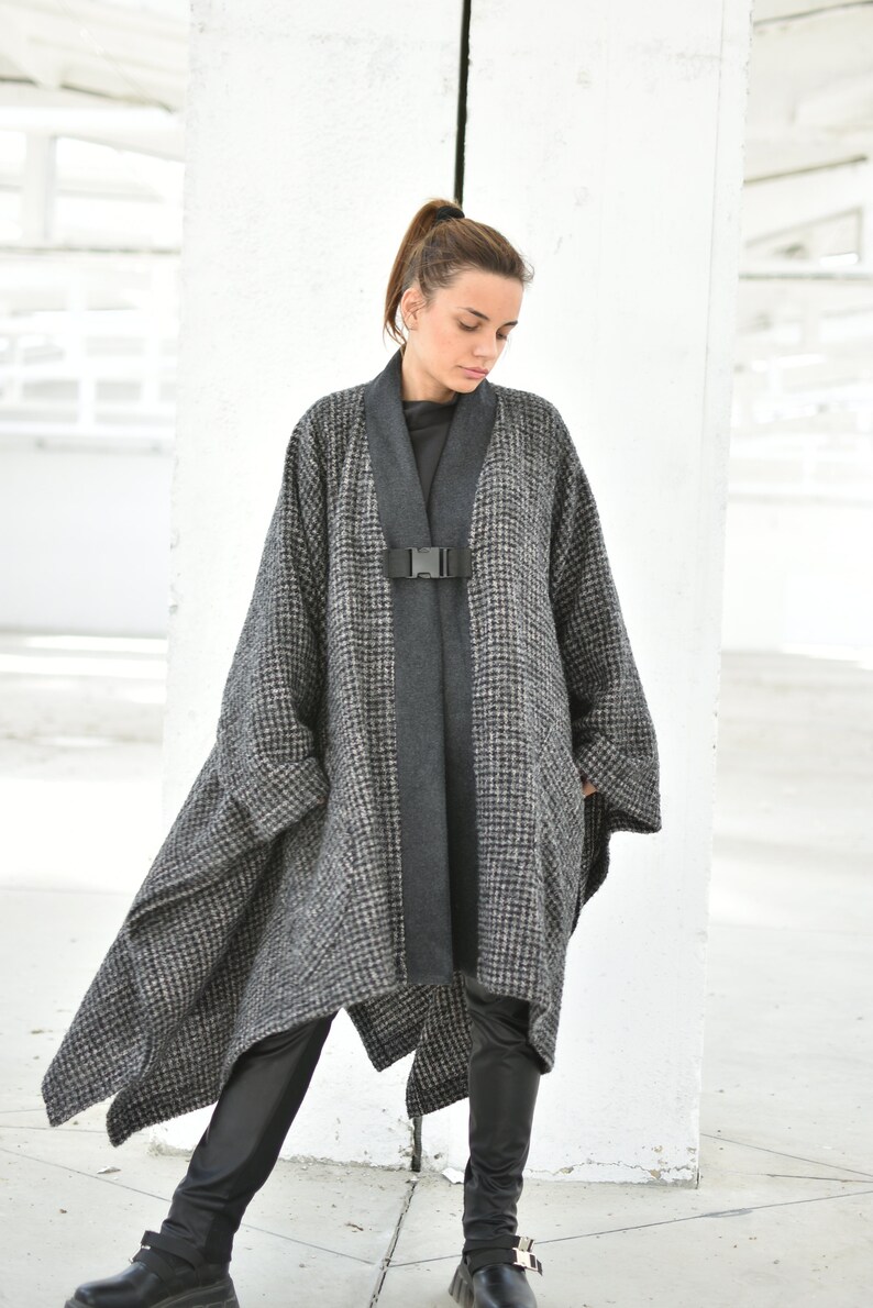 Pu&ograve; includere: Un poncho a motivi grigi e neri con una chiusura a fibbia nera. Il poncho ha un orlo lungo e asimmetrico ed &egrave; indossato sopra pantaloni neri.