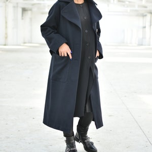 May include: A long navy blue coat with a large collar and pockets. The coat is worn over a black dress and black leggings. The person is wearing black boots with buckles.