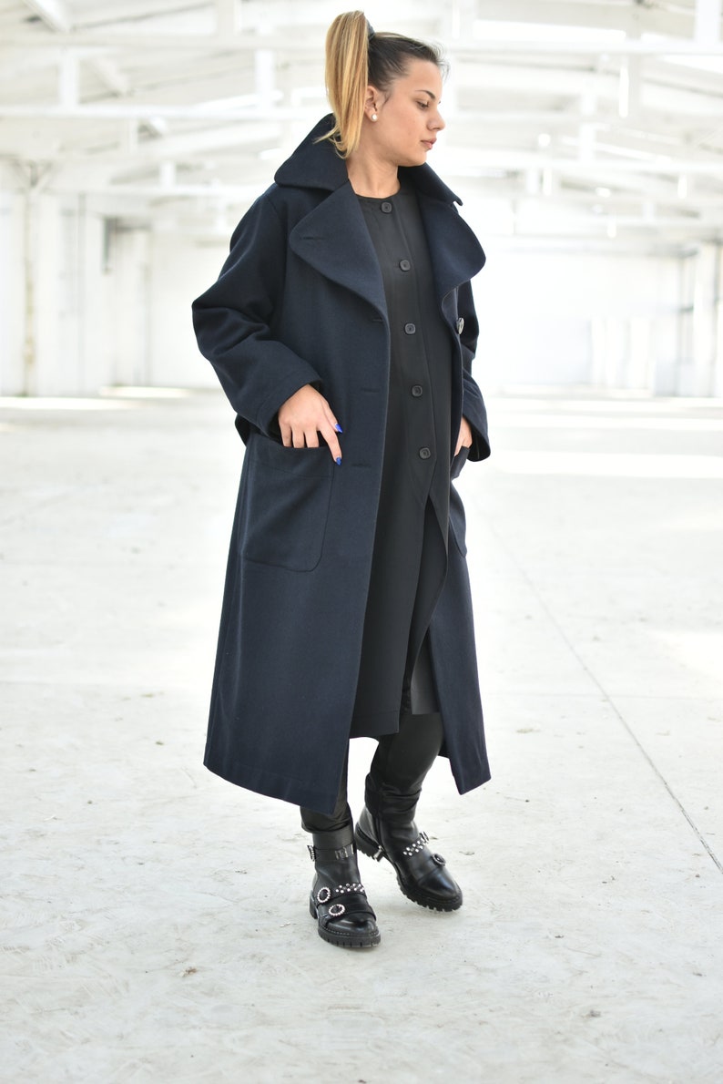 May include: A long navy blue coat with a large collar and pockets. The coat is worn over a black dress and black leggings. The person is wearing black boots with buckles.