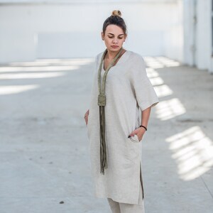 May include: A woman wearing a light beige linen dress and pants set with a long green fringe necklace. The outfit is a relaxed and comfortable style.