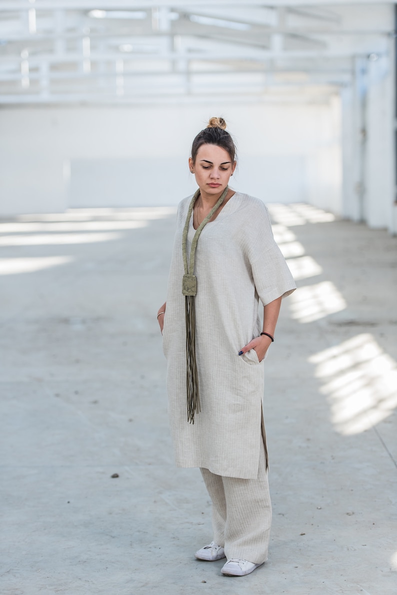 May include: A woman wearing a light beige linen dress and pants set with a long green fringe necklace. The outfit is a relaxed and comfortable style.