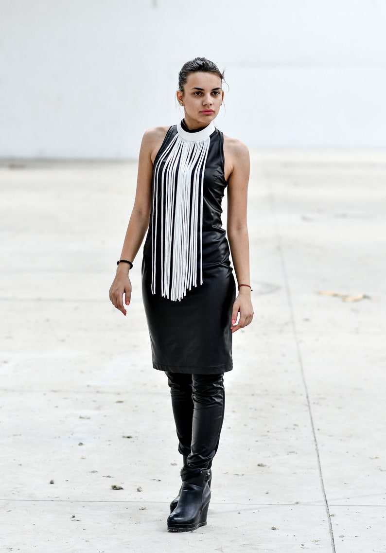 May include: A woman wearing a black leather dress with a white fringe detail at the neckline. She is wearing black leather boots and has her hair pulled back.
