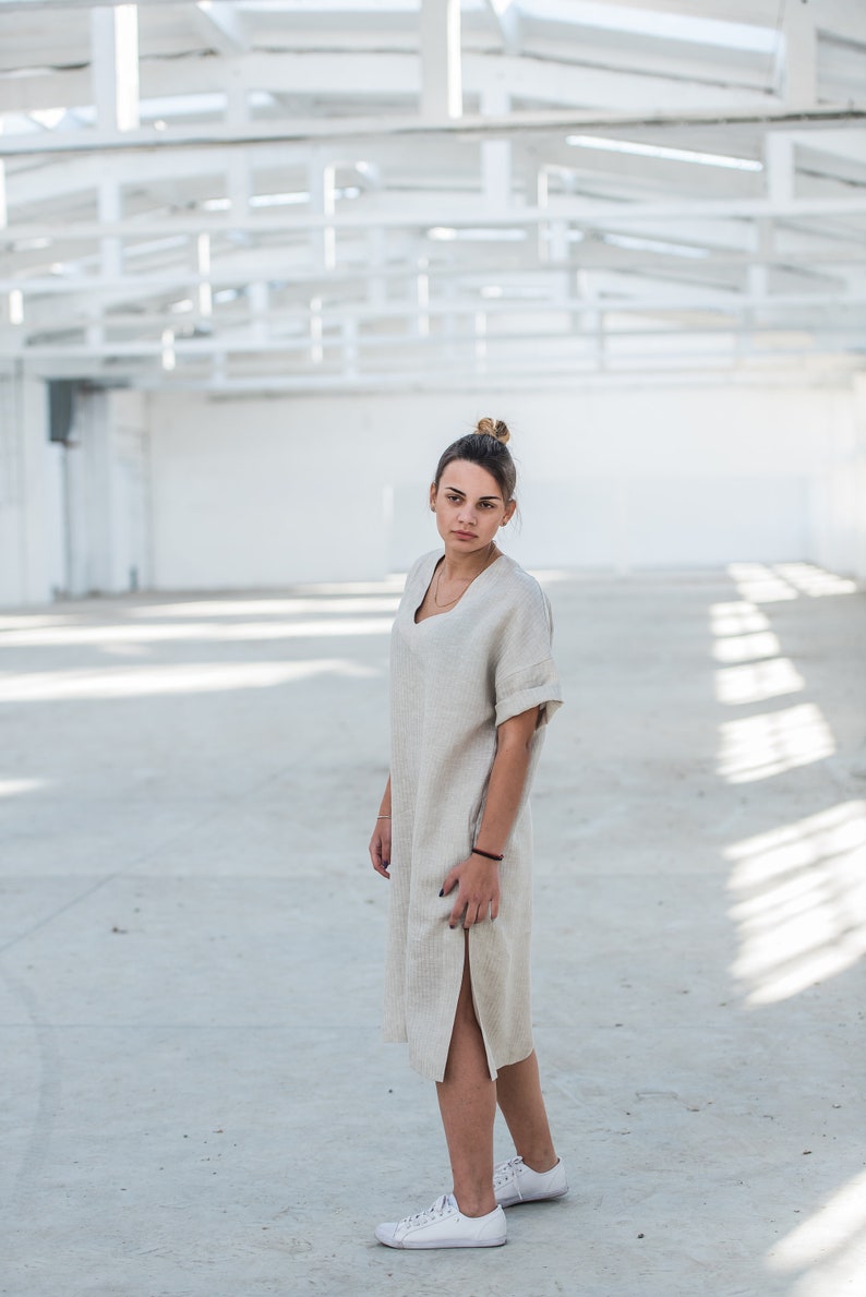 May include: A woman wearing a light beige linen dress with a side slit and white sneakers.