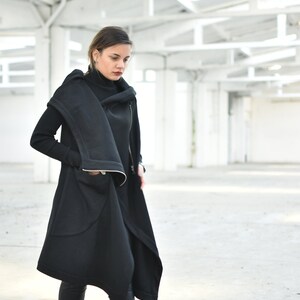 May include: A black, asymmetrical coat with a high collar and a long, flowing design. The coat features a zipper closure and a unique, draped silhouette. The person is wearing black leggings and boots. The background is a bright, open space with white walls and ceilings.