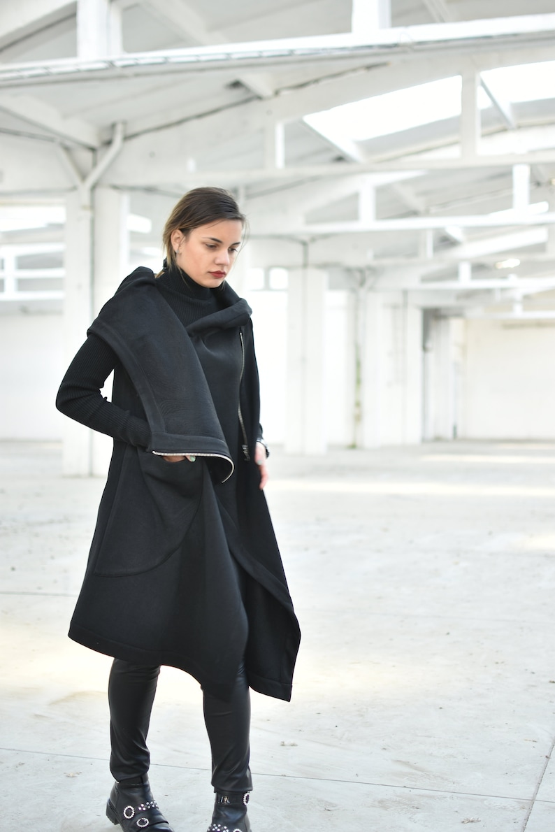 May include: A black, asymmetrical coat with a high collar and a long, flowing design. The coat features a zipper closure and a unique, draped silhouette. The person is wearing black leggings and boots. The background is a bright, open space with white walls and ceilings.