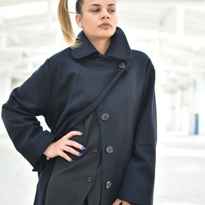 May include: A woman wearing a long, dark blue coat with a high collar and multiple buttons. The coat is styled with a wrap-over design and has a relaxed fit.