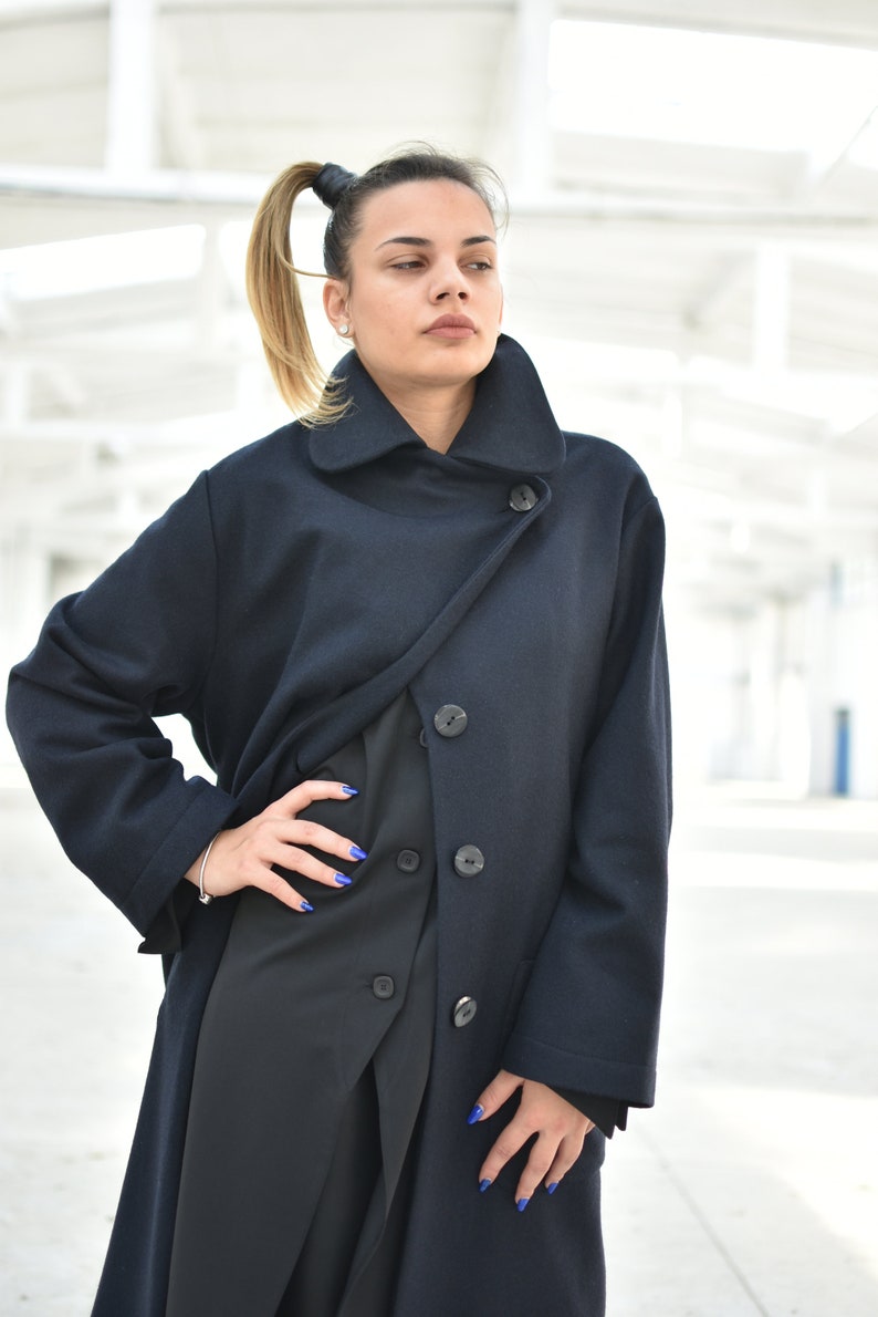 May include: A woman wearing a long, dark blue coat with a high collar and multiple buttons. The coat is styled with a wrap-over design and has a relaxed fit.