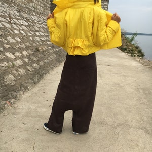 May include: A bright yellow puffer jacket with a ruffled hem is worn over a pair of dark brown, wide-legged pants. The person is wearing black and white sneakers. The outfit is outdoors on a concrete path.