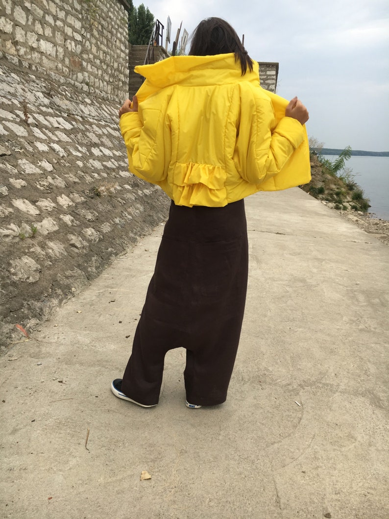 May include: A bright yellow puffer jacket with a ruffled hem is worn over a pair of dark brown, wide-legged pants. The person is wearing black and white sneakers. The outfit is outdoors on a concrete path.