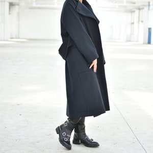 May include: A woman wearing a long, dark blue coat with a high collar and a pair of black boots with buckles.