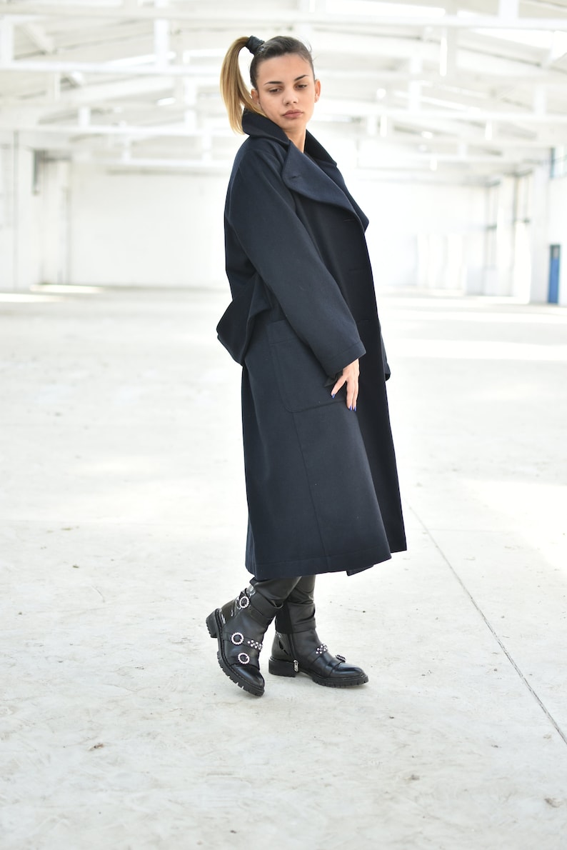 May include: A woman wearing a long, dark blue coat with a high collar and a pair of black boots with buckles.