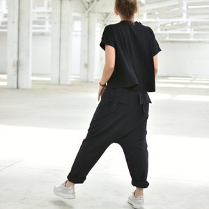 May include: A person wearing a black short-sleeved shirt and black pants with a unique design. The person is standing in a large, white, empty space.
