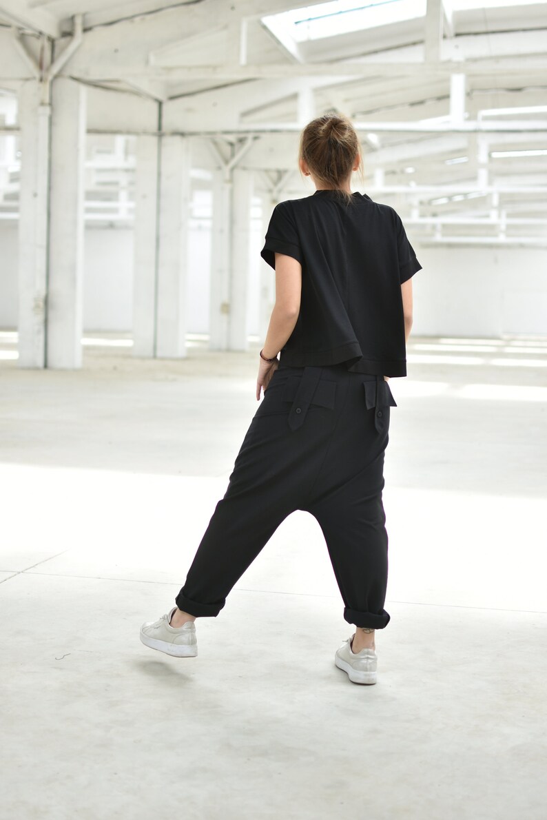 May include: A person wearing a black short-sleeved shirt and black pants with a unique design. The person is standing in a large, white, empty space.