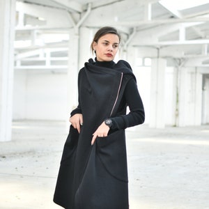 May include: A long black coat with a unique asymmetrical design. The coat features a high collar and a pink zipper detail. The person is wearing a black turtleneck and a watch. The coat has large pockets and a flowing silhouette.