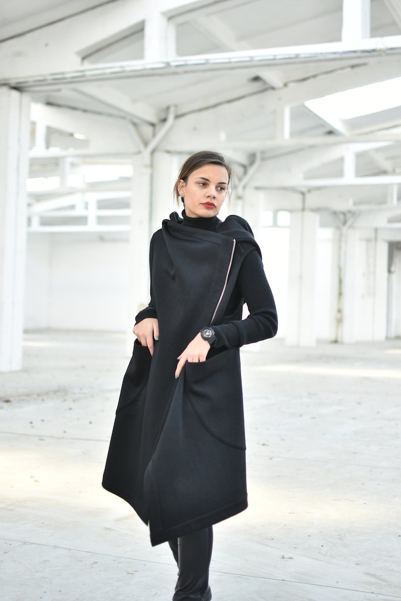 May include: A long black coat with a unique asymmetrical design. The coat features a high collar and a pink zipper detail. The person is wearing a black turtleneck and a watch. The coat has large pockets and a flowing silhouette.