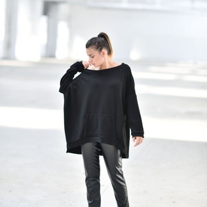 May include: A woman wearing a black oversized sweater with a pocket and black leather pants. She is standing in a white, industrial setting.
