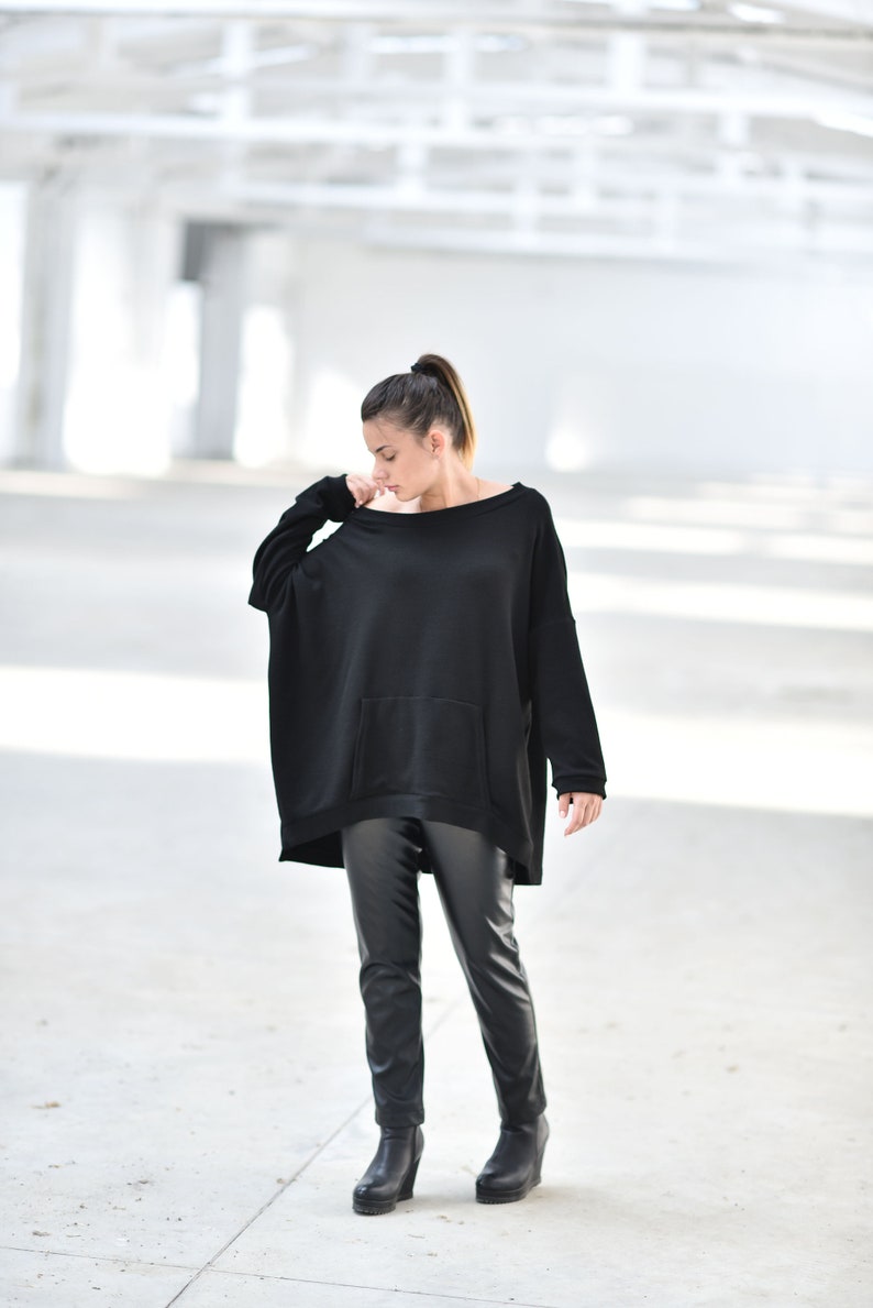 May include: A woman wearing a black oversized sweater with a pocket and black leather pants. She is standing in a white, industrial setting.