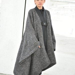 Pu&ograve; includere: Un lungo cappotto a quadri bianco e nero con una cintura nera e fibbia. Il cappotto &egrave; asimmetrico e ha una vestibilit&agrave; ampia. La modella indossa pantaloni neri e scarpe nere.