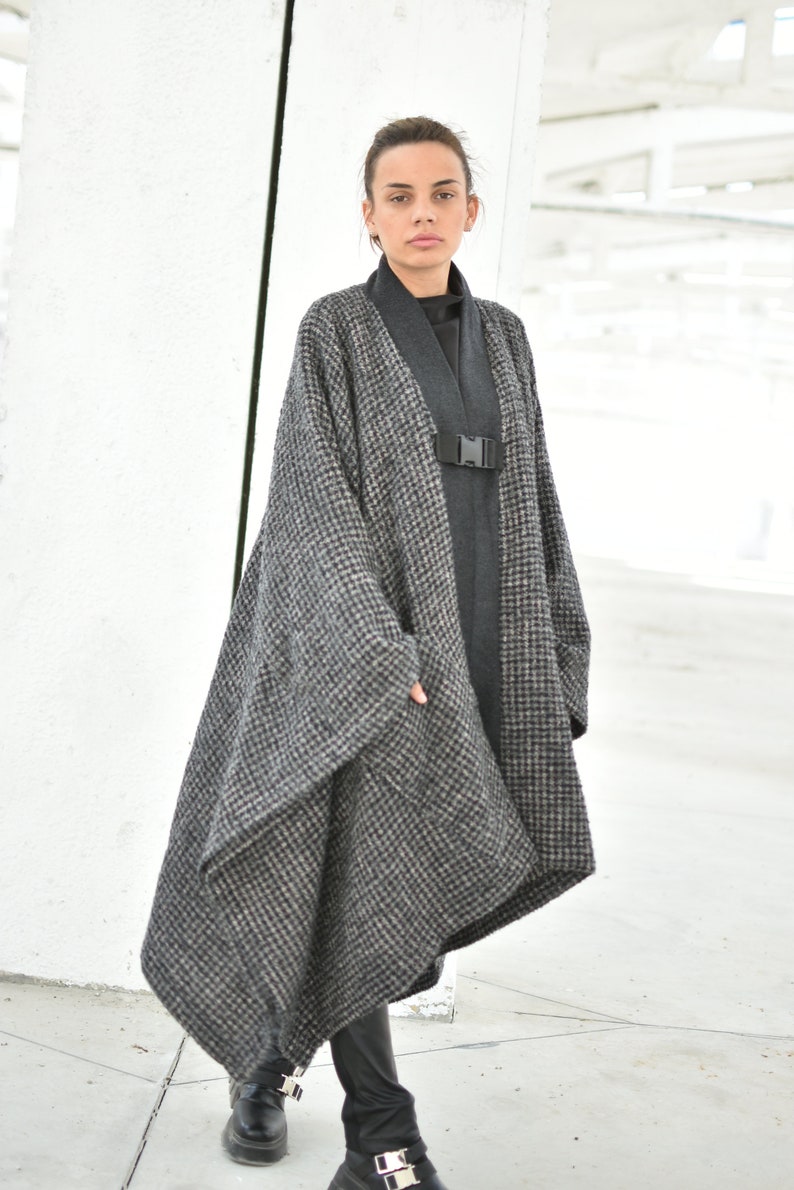 Pu&ograve; includere: Un lungo cappotto a quadri bianco e nero con una cintura nera e fibbia. Il cappotto &egrave; asimmetrico e ha una vestibilit&agrave; ampia. La modella indossa pantaloni neri e scarpe nere.