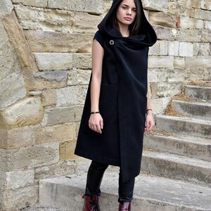May include: A long black sleeveless hooded vest with a draped collar and a single button closure. The vest is worn over black leggings and red leather boots.