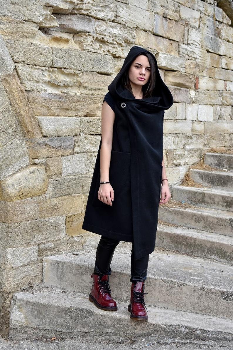 May include: A long black sleeveless hooded vest with a draped collar and a single button closure. The vest is worn over black leggings and red leather boots.