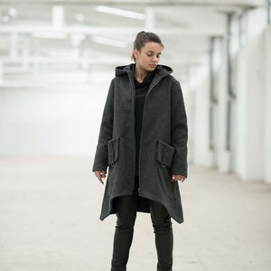 May include: A woman wearing a long, gray hooded coat with a zipper closure and two pockets. She is wearing black pants and black boots.