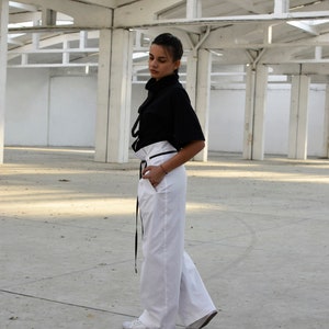 May include: A woman wearing a black mock neck shirt with a white high-waisted pant and black belt. She is standing in a white room with a concrete floor.
