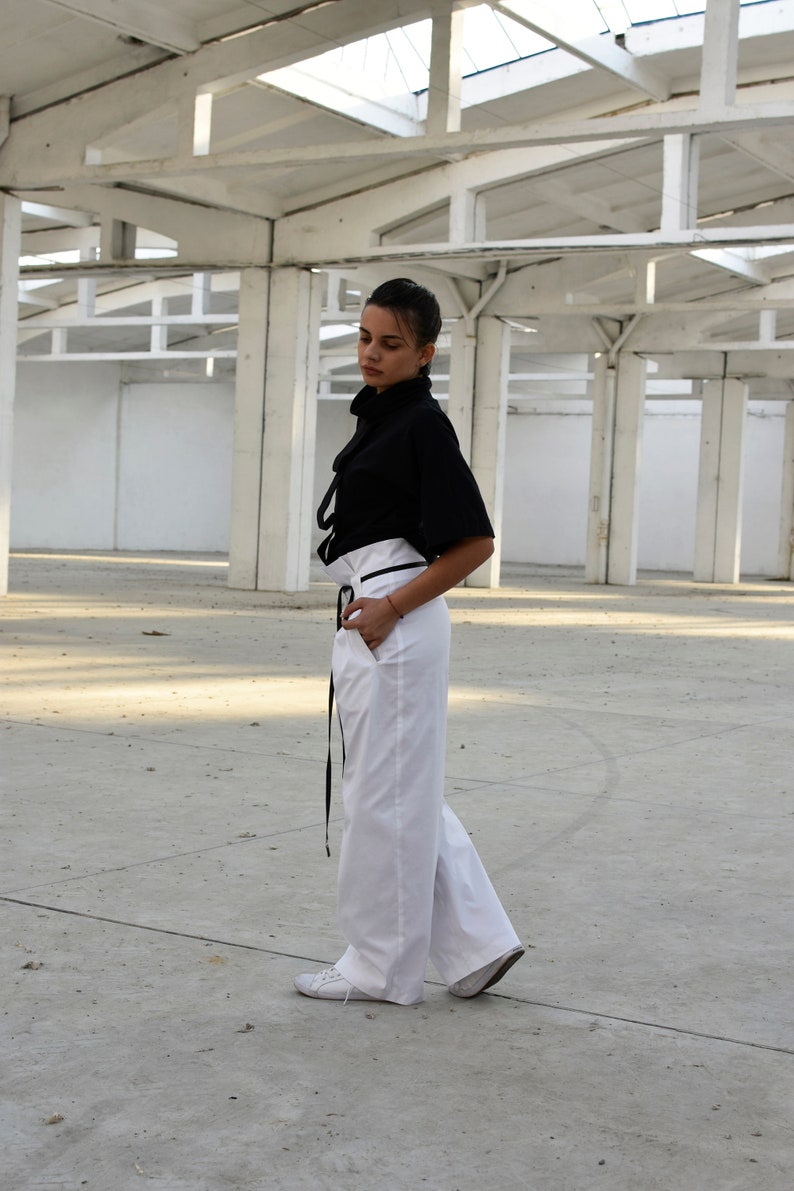 May include: A woman wearing a black mock neck shirt with a white high-waisted pant and black belt. She is standing in a white room with a concrete floor.