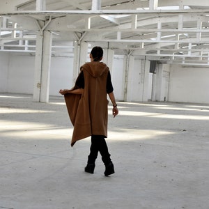 May include: A person wearing a brown hooded vest with a long, flowing design. The vest has a relaxed fit and is worn over a black shirt. The person is standing in a large, empty room with a white ceiling.